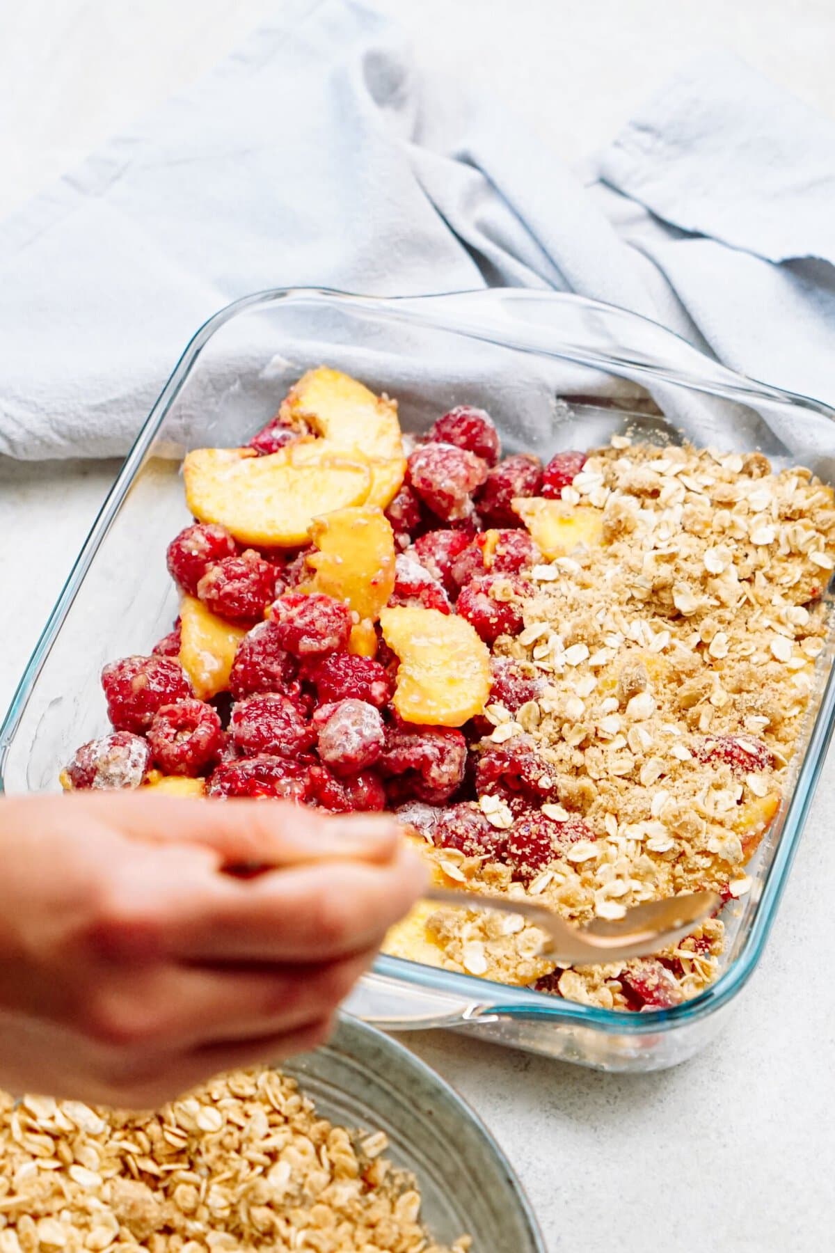 A hand is sprinkling oat crumble over a mixture of frozen raspberries and peach slices in a glass baking dish, preparing a delightful raspberry cobbler. A light blue cloth lies softly in the background.