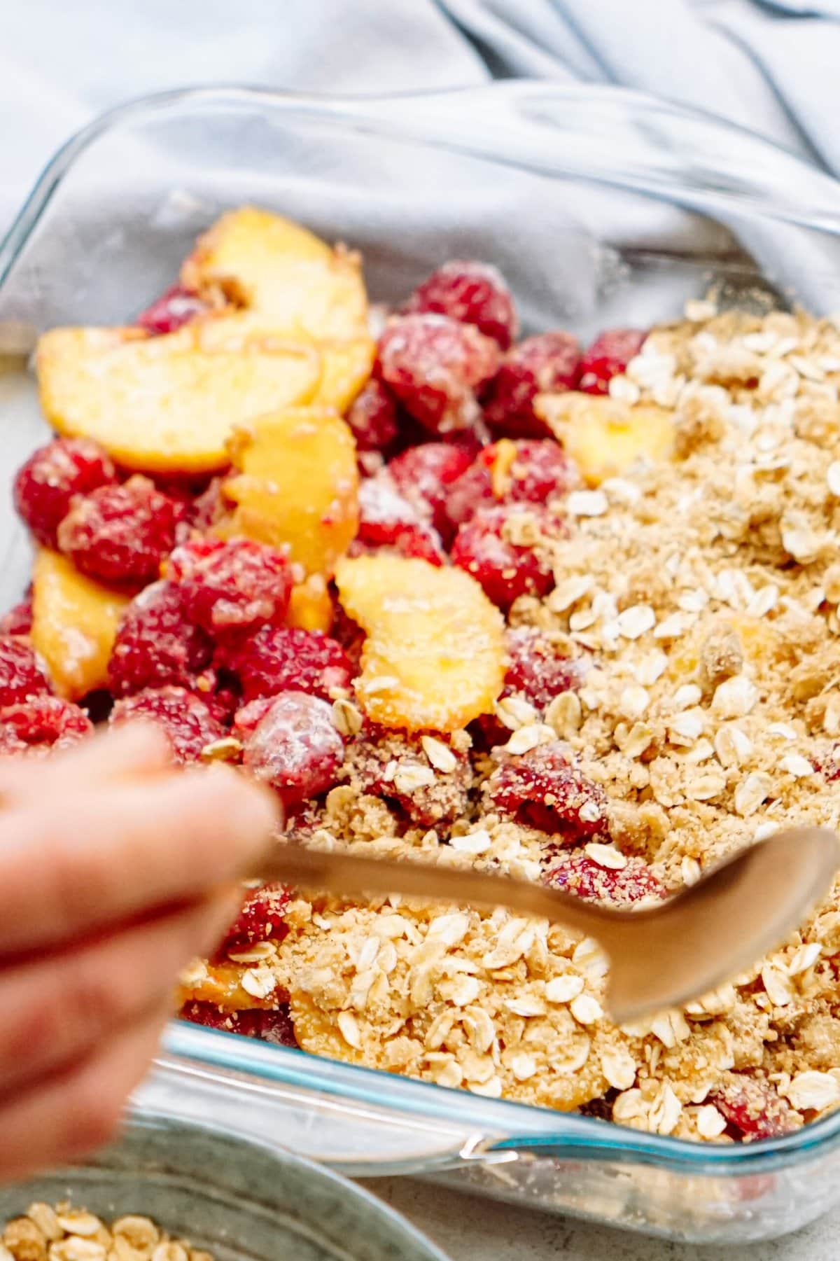 A hand holding a spoon is shown near a glass baking dish containing peach and raspberry cobbler topped with oats and a crumbly brown sugar mixture.