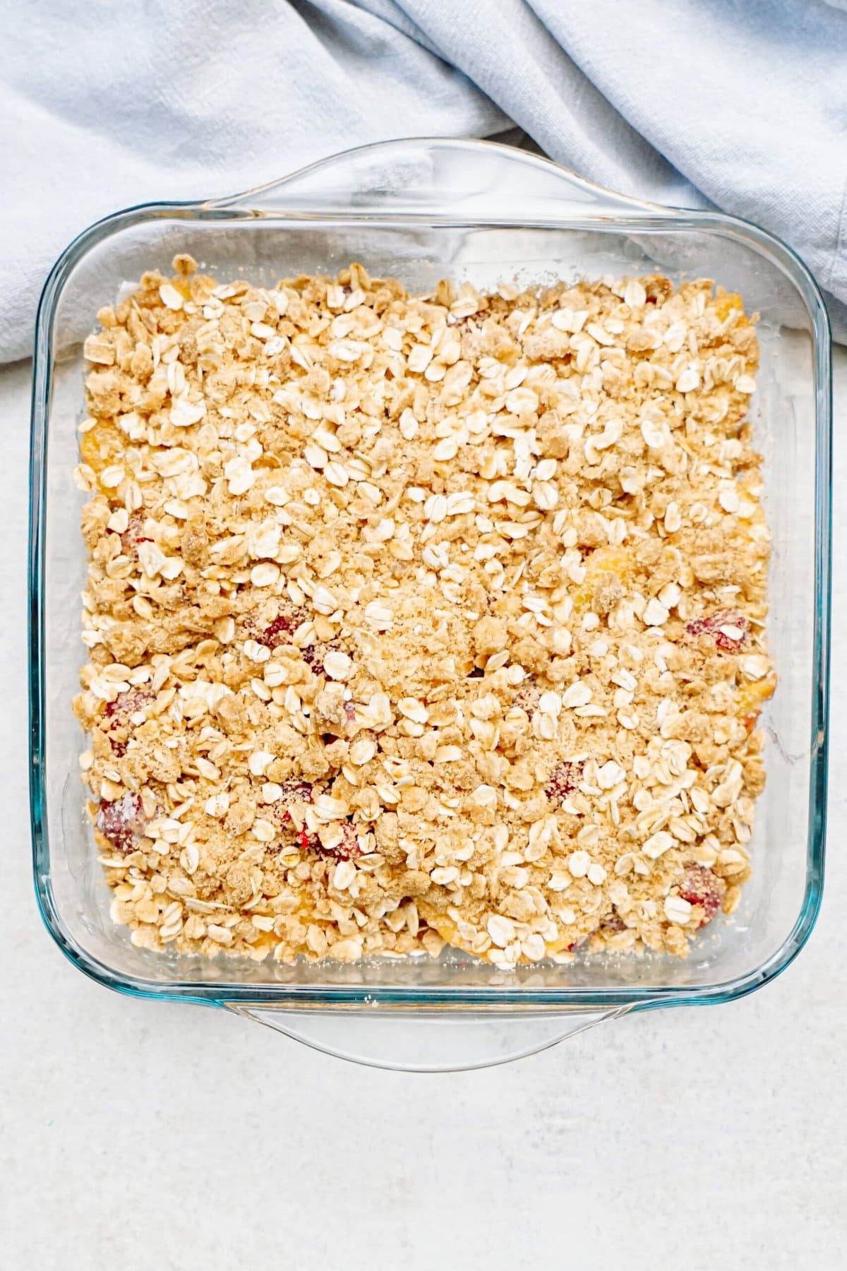 A glass baking dish filled with an unbaked oatmeal raspberry cobbler mixture is set on a light surface with a blue cloth nearby.