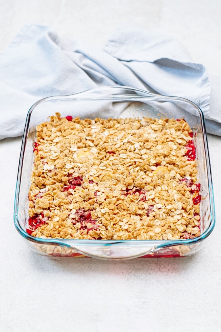 A glass baking dish with strawberry and raspberry cobbler topped with oats and crumble mixture, sitting on a light-colored surface next to a folded cloth.