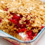 A serving of raspberry cobbler with an oat topping is being scooped from a glass baking dish.
