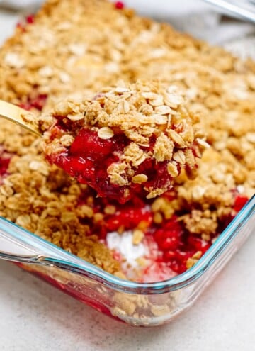 A serving of raspberry cobbler with an oat topping is being scooped from a glass baking dish.