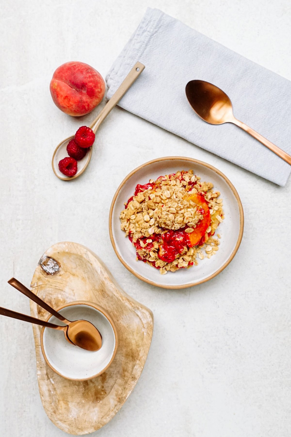A plate of raspberry cobbler next to a spoon, fresh peach, and raspberries. A wooden tray holds a bowl and spoons. A folded napkin with a spoon is placed nearby on a light surface.