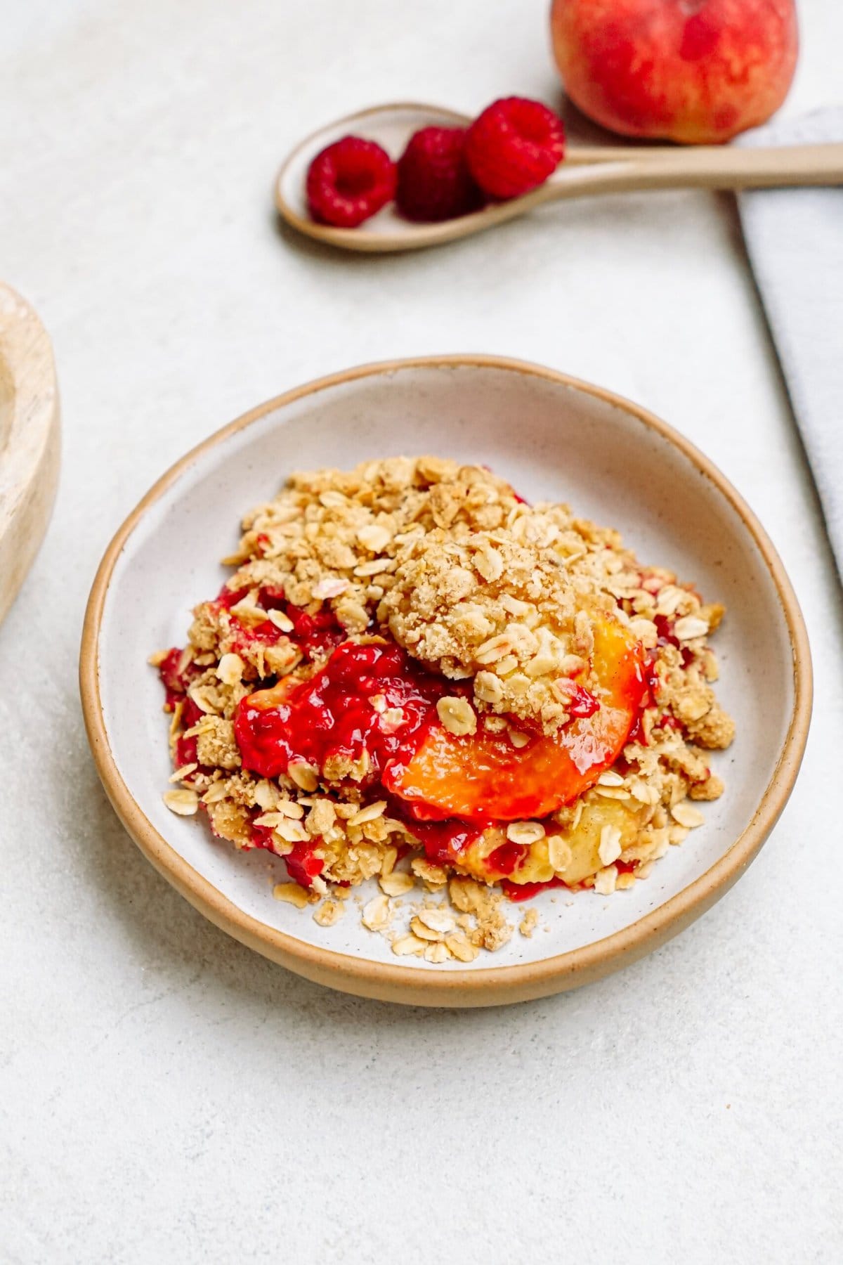 A bowl of raspberry cobbler topped with a crumbly oat mixture on a white plate. A wooden spoon with raspberries and a peach are in the background.