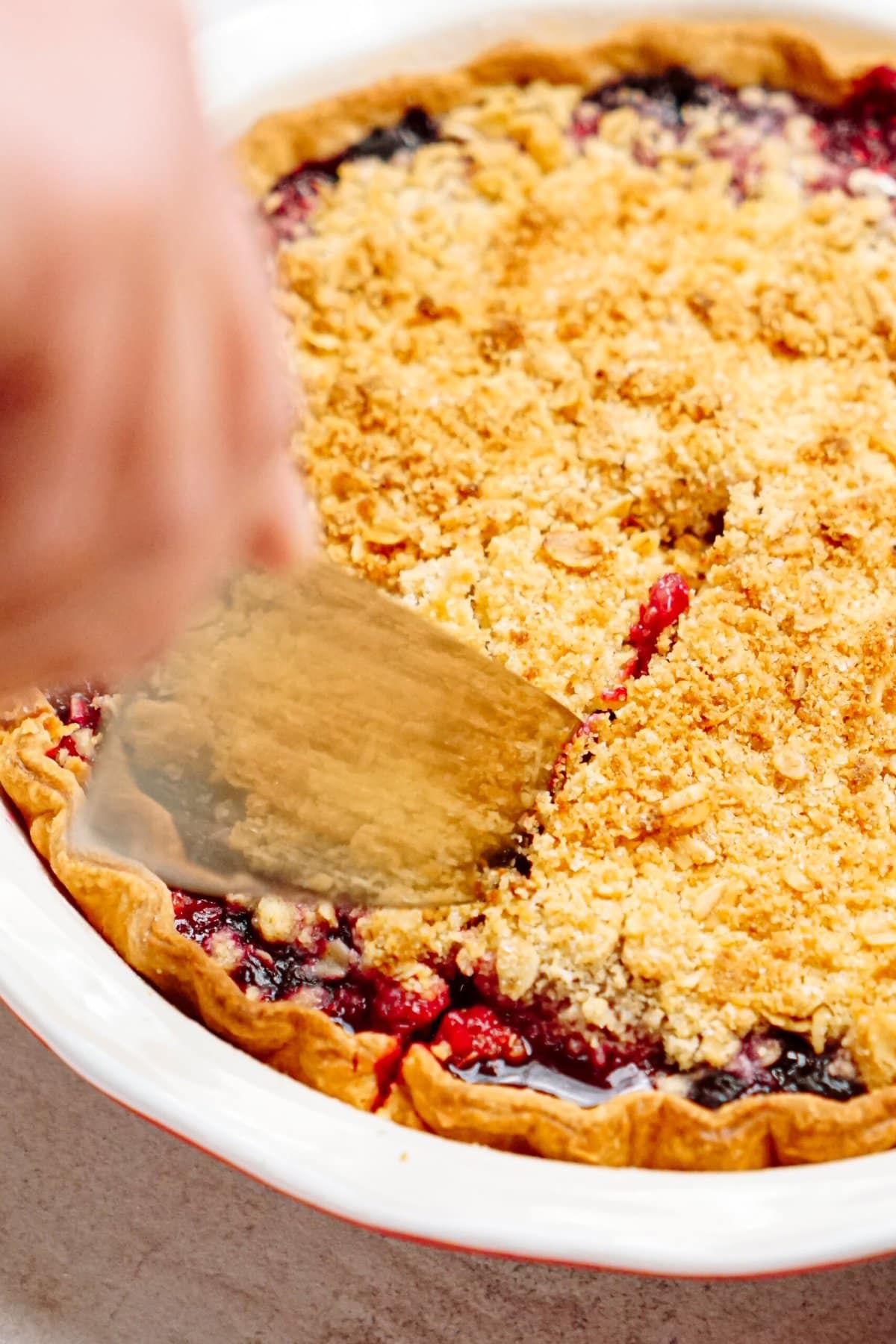 A hand using a metal spatula to slice into a berry crumble pie with a golden crumb topping.