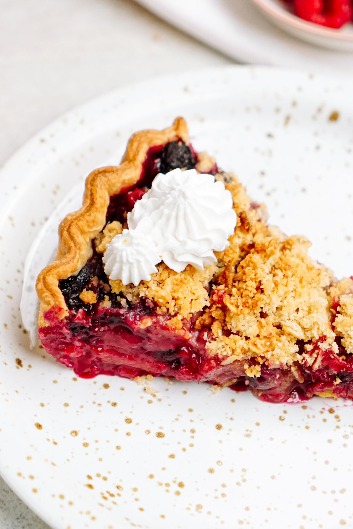 A slice of berry crumble pie topped with a dollop of whipped cream on a white plate.