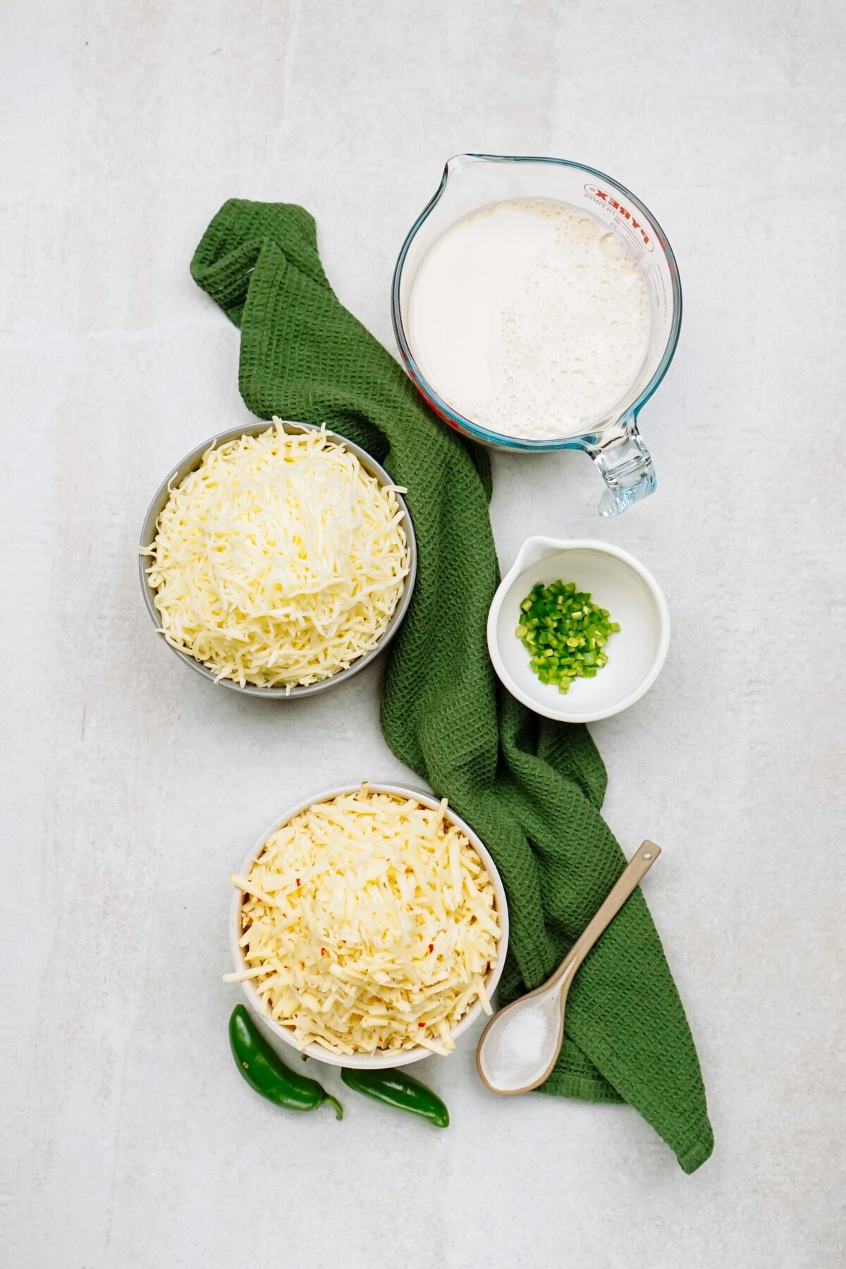 Top-down view of shredded cheese in two bowls, a small bowl of chopped green peppers, a measuring cup of milk for the queso dip, a wooden spoon with salt, and green cloth on a light surface.