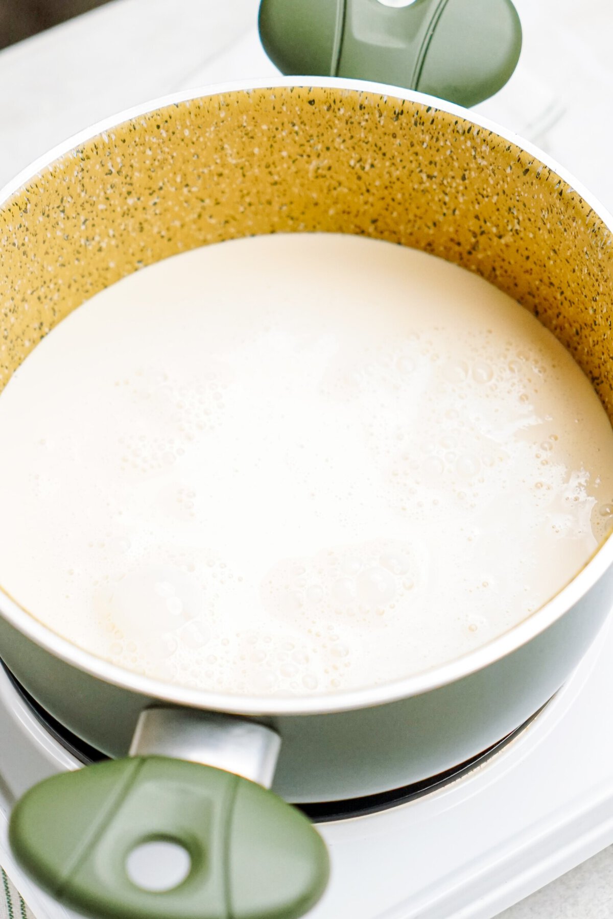 A green cooking pot with a speckled yellow interior contains a bubbling white queso dip, placed on a white stovetop.