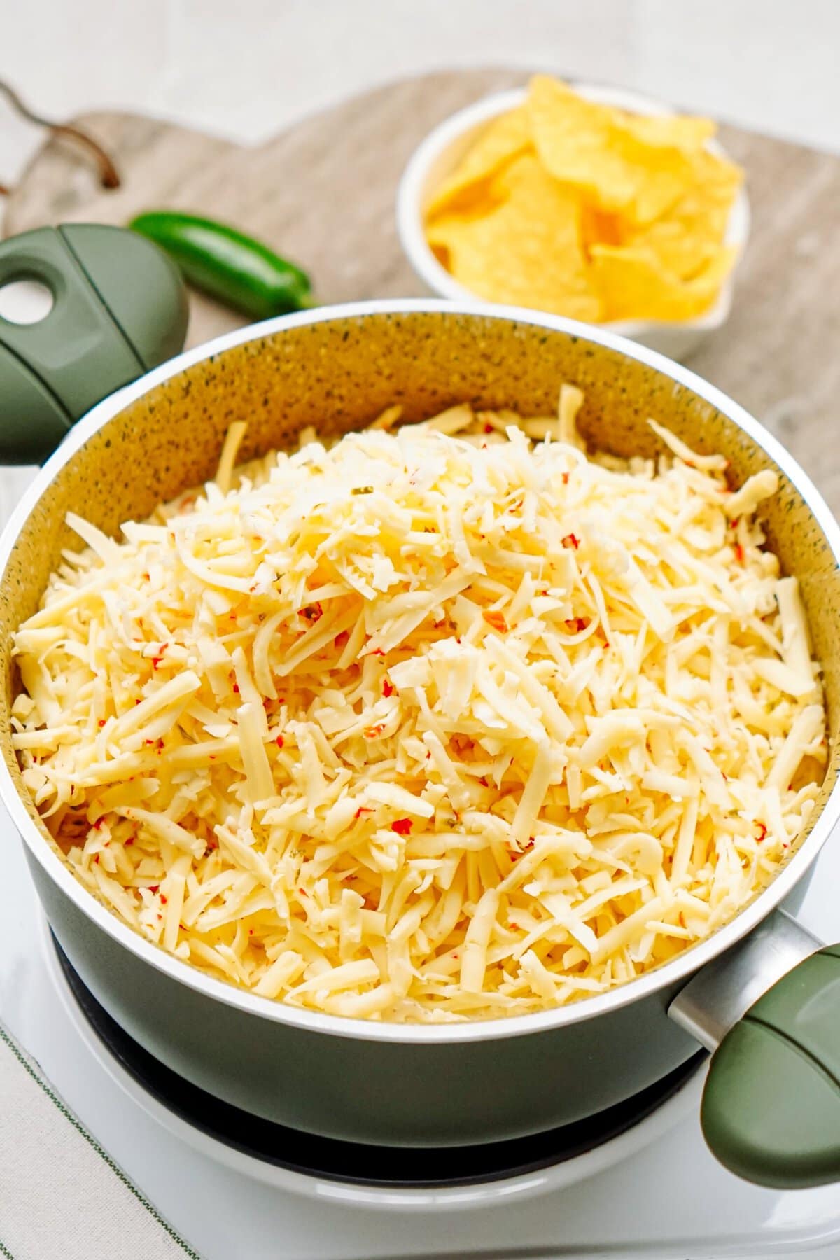 A pot filled with shredded cheese, ready to become a delicious queso dip, is placed on an electric stovetop. In the background, a bowl of tortilla chips is slightly blurred.