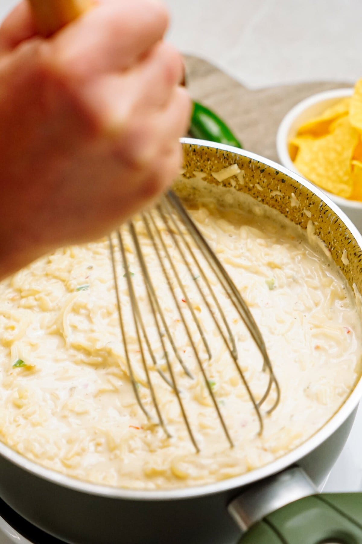 A hand is whisking creamy white sauce in a pot on the stove, transforming it into a savory queso dip. A bowl of yellow chips is visible in the background, ready to be paired with the delicious dip.