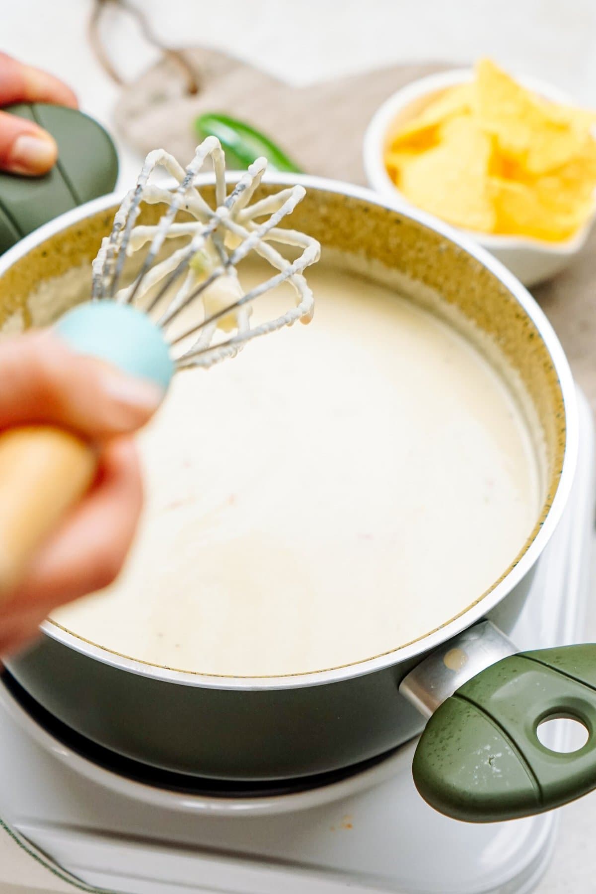 Hand whisking creamy queso dip in a green pot on a stove, with a small bowl of yellow tortilla chips in the background.
