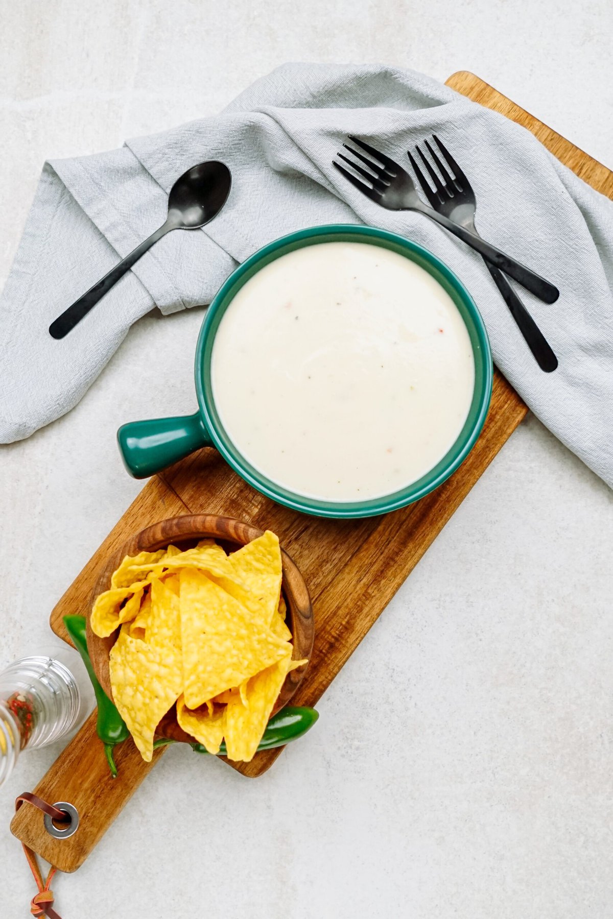 A bowl of creamy white queso dip with a side of tortilla chips is on a wooden serving board. Nearby are two black forks, a black spoon, and a gray napkin.