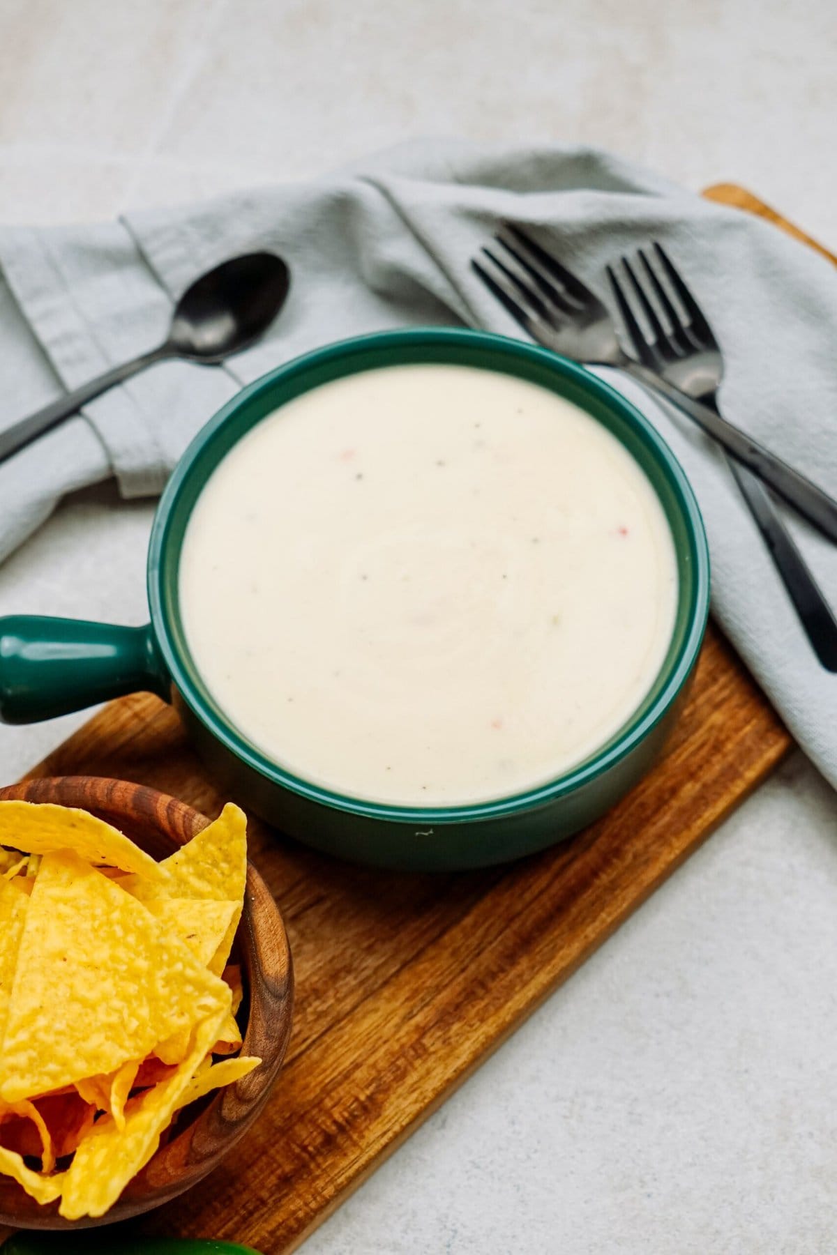 A green bowl filled with creamy white queso dip sits on a wooden board, accompanied by a small bowl of tortilla chips. Silverware and a folded napkin rest in the background, completing this inviting scene.