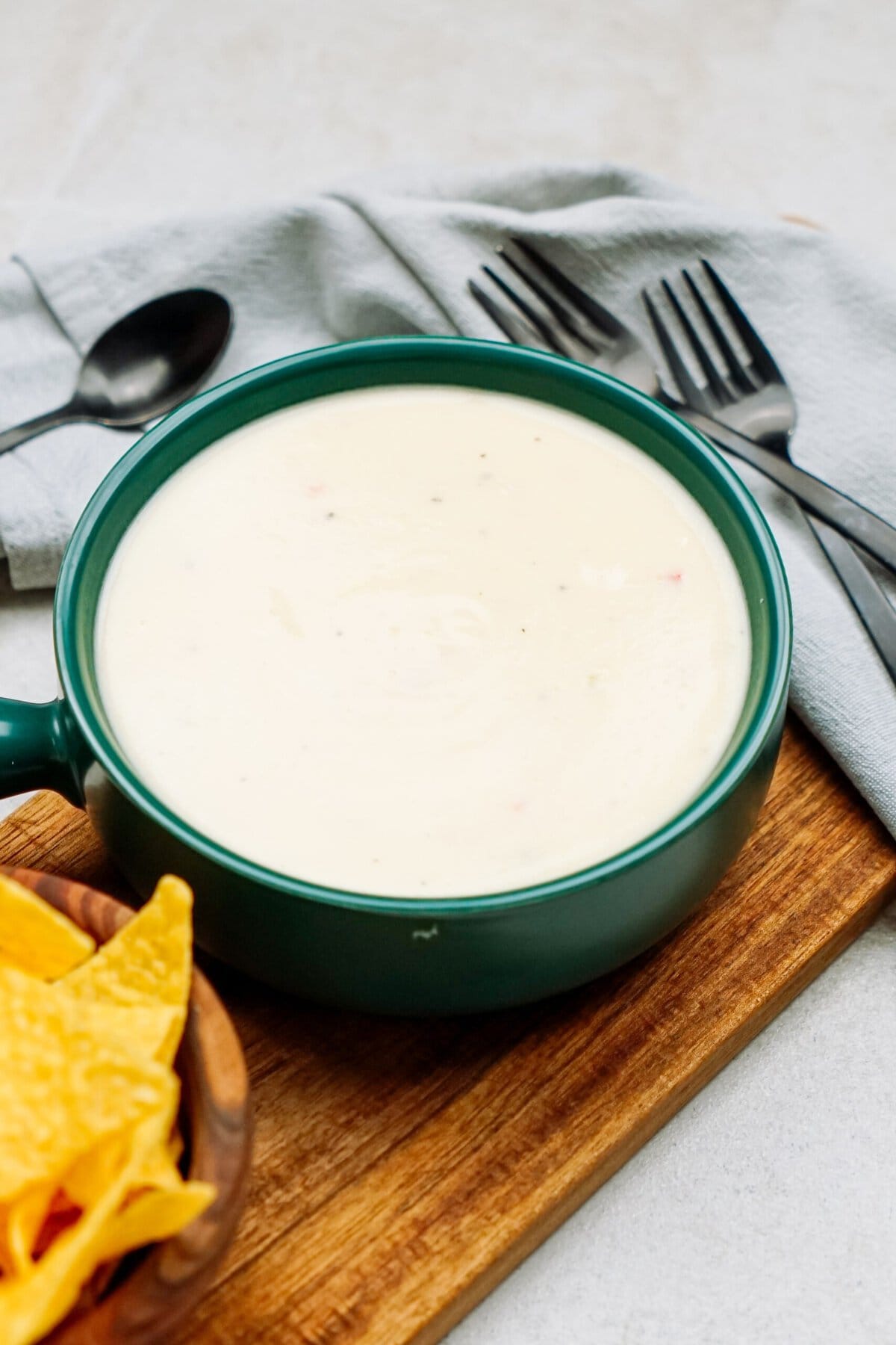 A bowl of creamy white queso dip on a wooden board next to a small bowl of tortilla chips. Silverware and a gray cloth napkin are placed in the background.