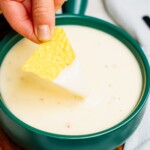 A hand dipping a triangular tortilla chip into a bowl of creamy queso dip placed on a wooden surface.