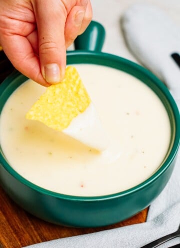 A hand dipping a triangular tortilla chip into a bowl of creamy queso dip placed on a wooden surface.