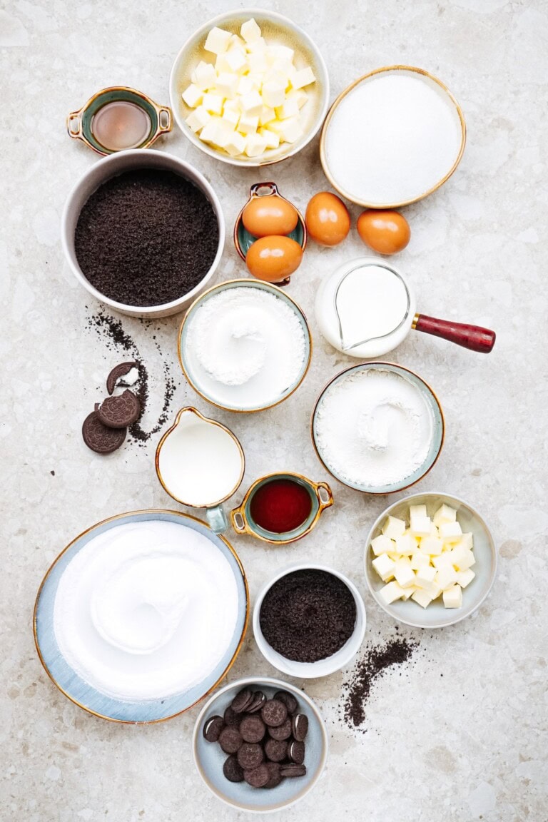 Top-down view of baking ingredients including butter, sugar, eggs, vanilla, flour, cocoa powder, small chocolate cookies, and a few other measured items on a light-colored countertop.