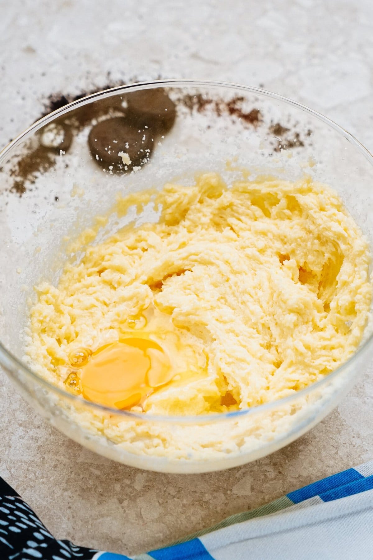 A glass bowl on a countertop containing creamed butter and sugar with an egg cracked in the center, ready to be mixed.