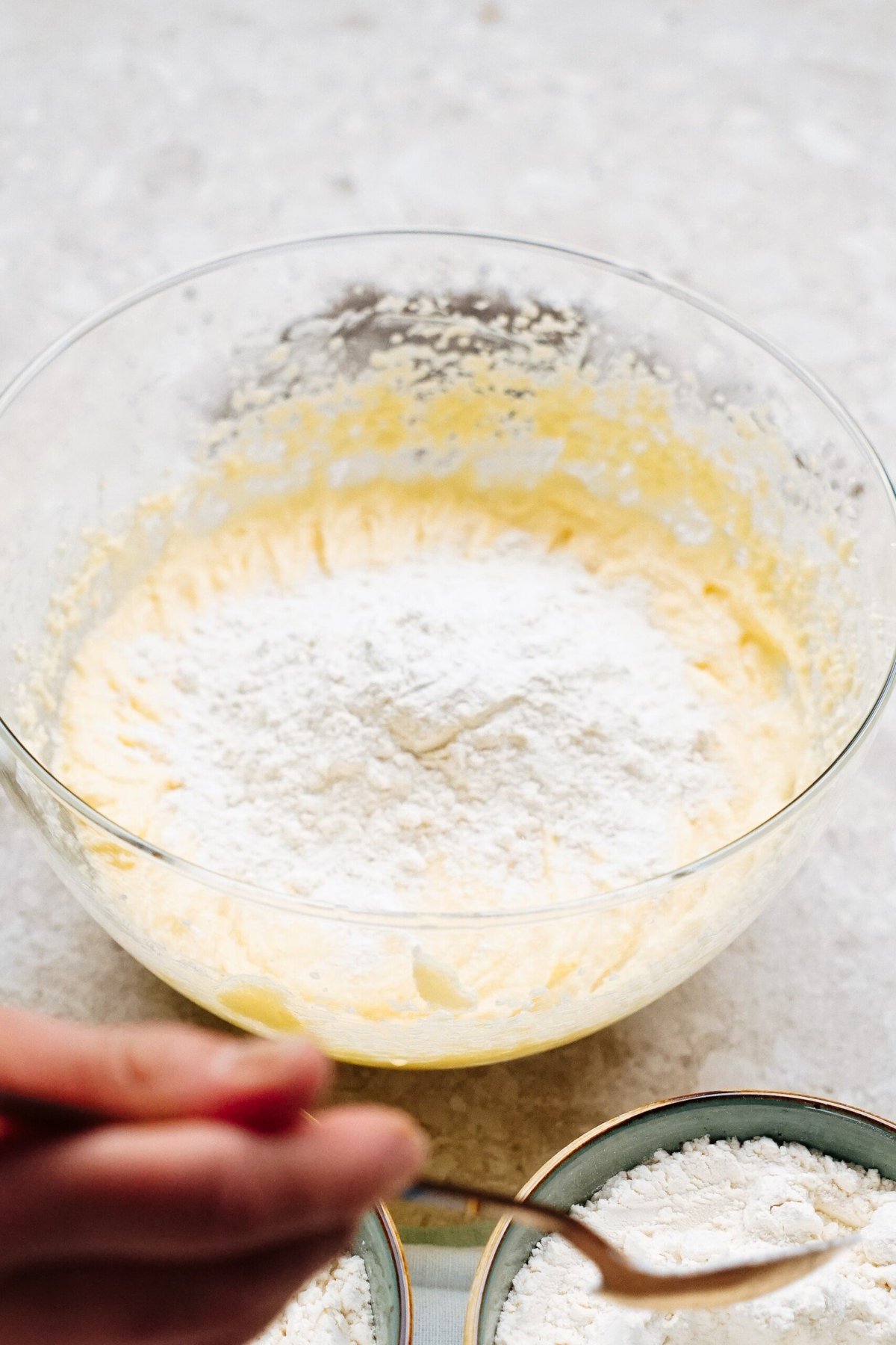 A bowl containing a mixture of creamed butter and sugar, with flour being added on top. A hand holding a spoon is visible in the foreground.