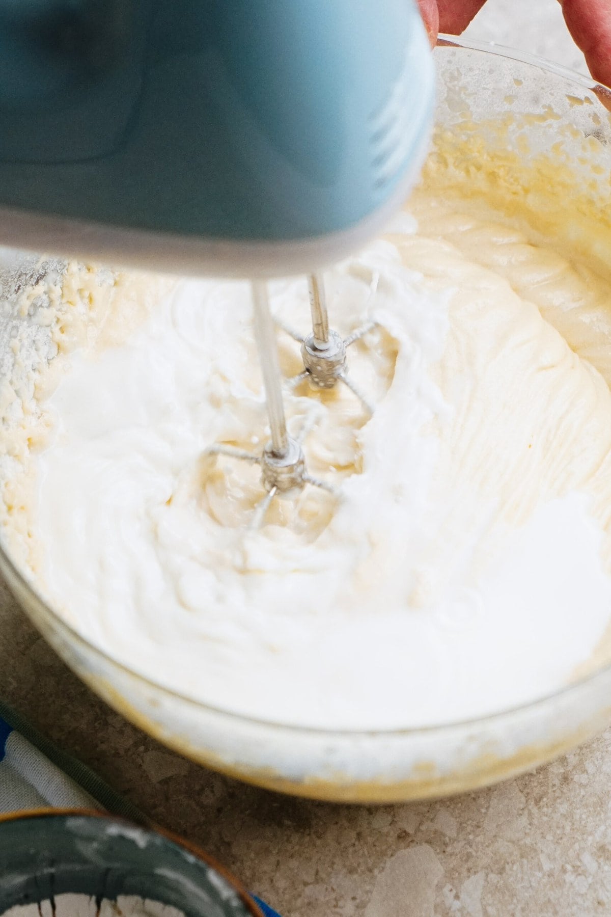 A blue hand mixer is being used to blend ingredients in a glass bowl filled with creamy batter.