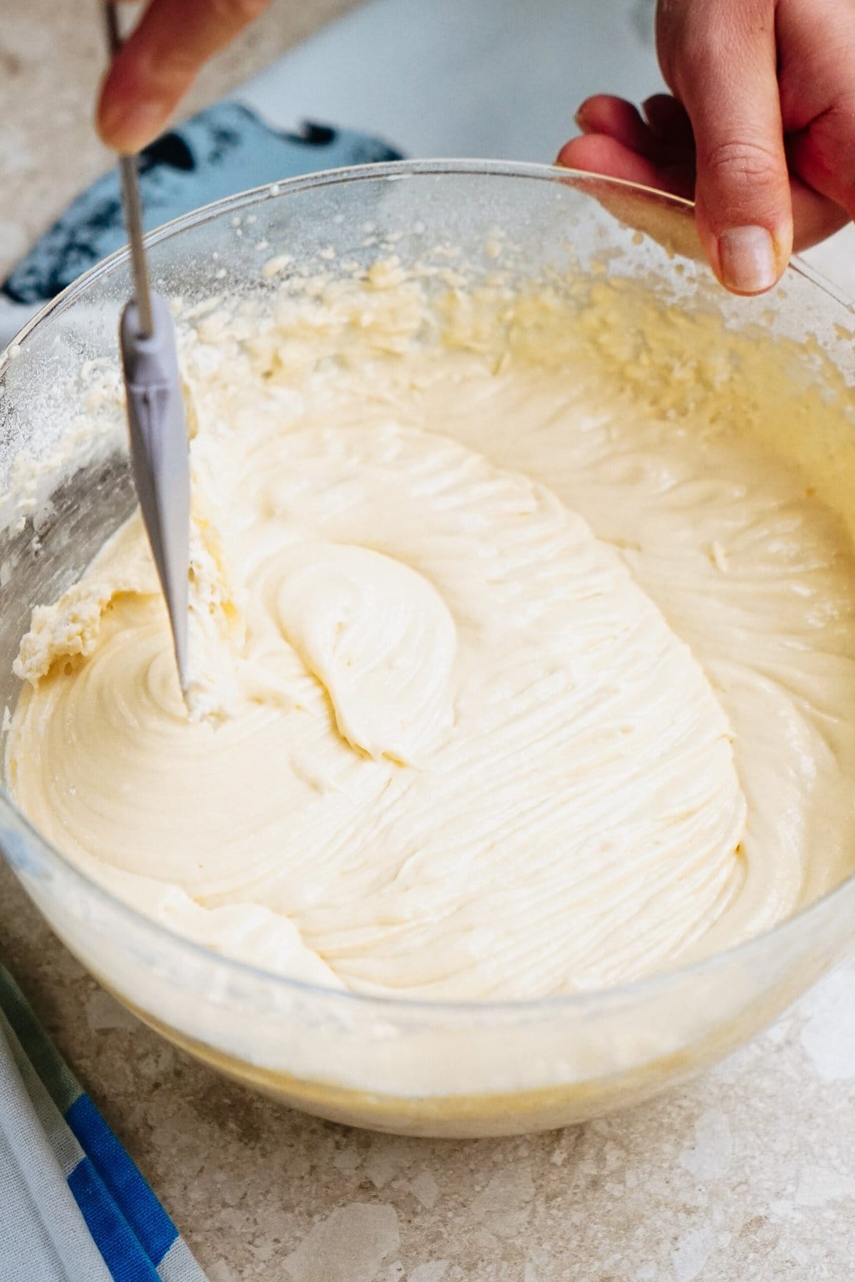 A hand is mixing smooth, creamy batter in a clear glass bowl with a metal whisk on a light-colored surface.