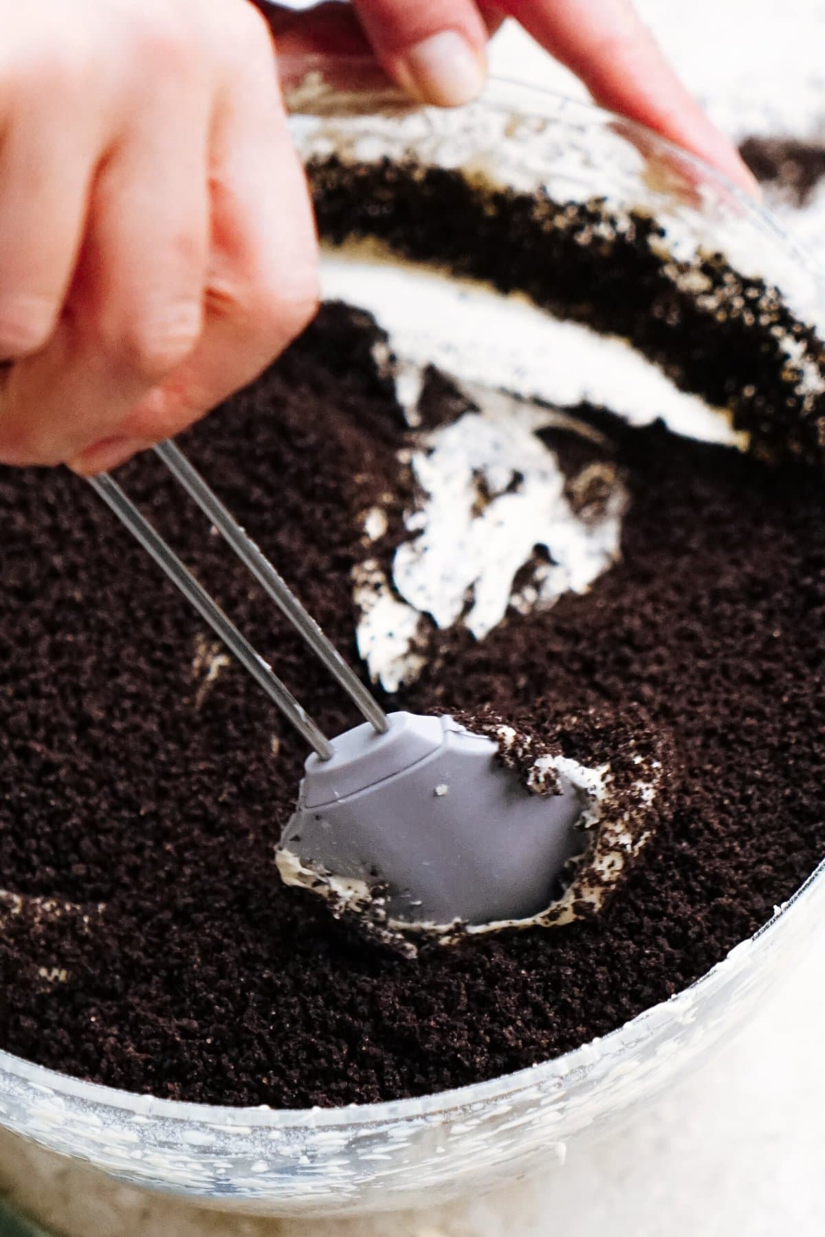 Close-up of a hand holding a spoon and scooping a mixture of crushed chocolate cookies and cream from a glass bowl.