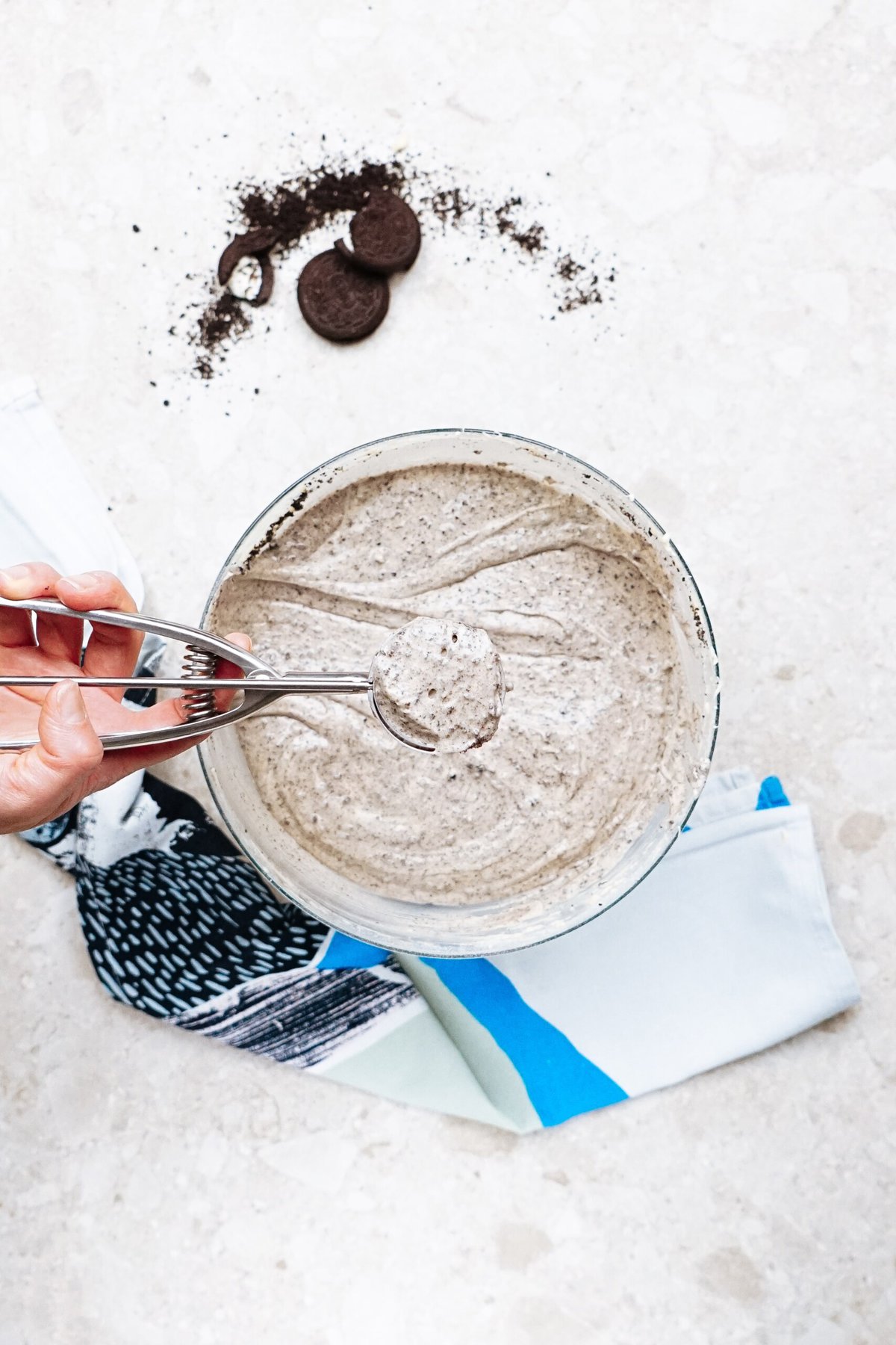 A hand using a scoop to take some cookies and cream batter from a bowl. Napkins and cookie crumbs are scattered on the surface around the bowl.