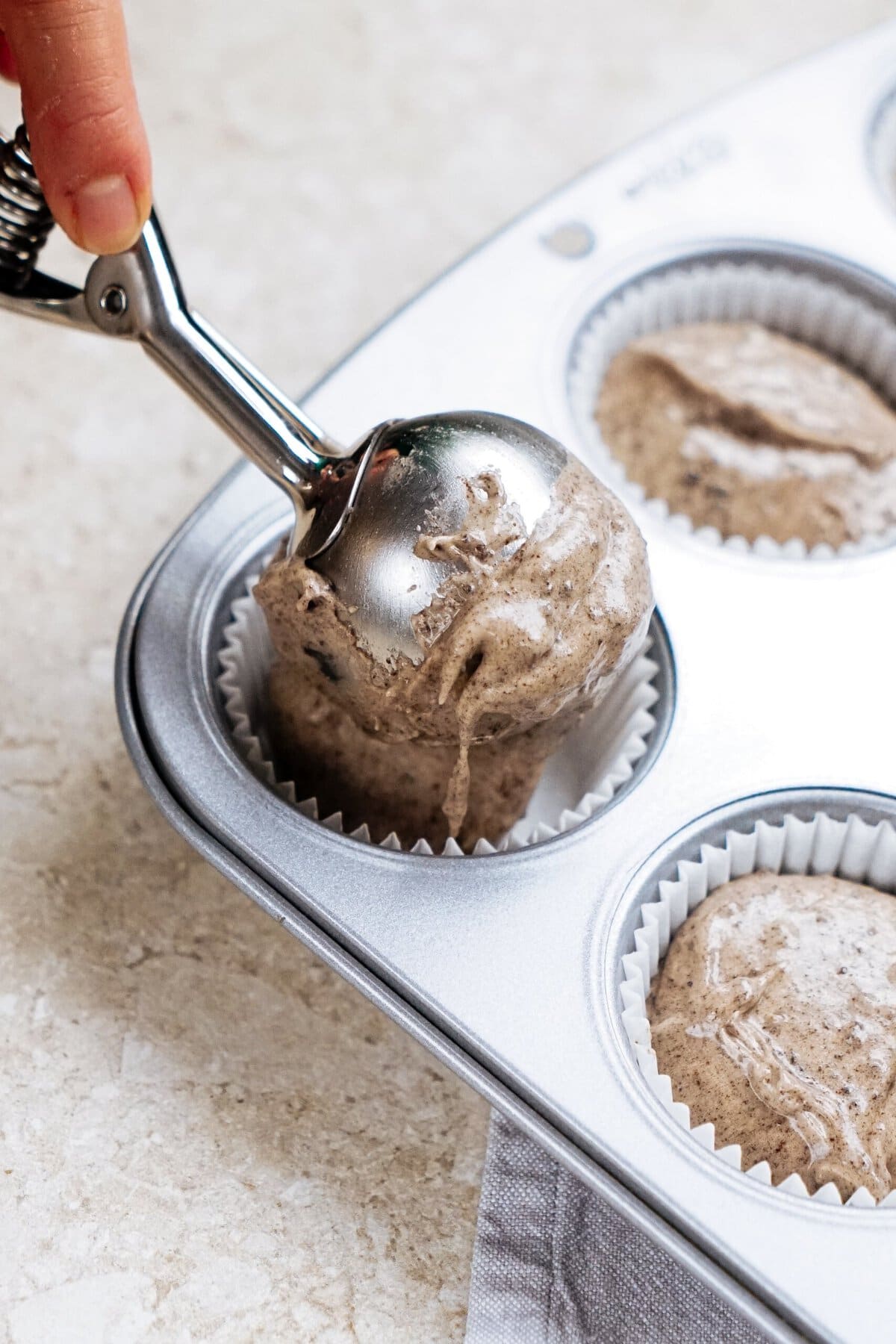 A metal scoop is used to place cupcake batter into white paper liners in a muffin tin.