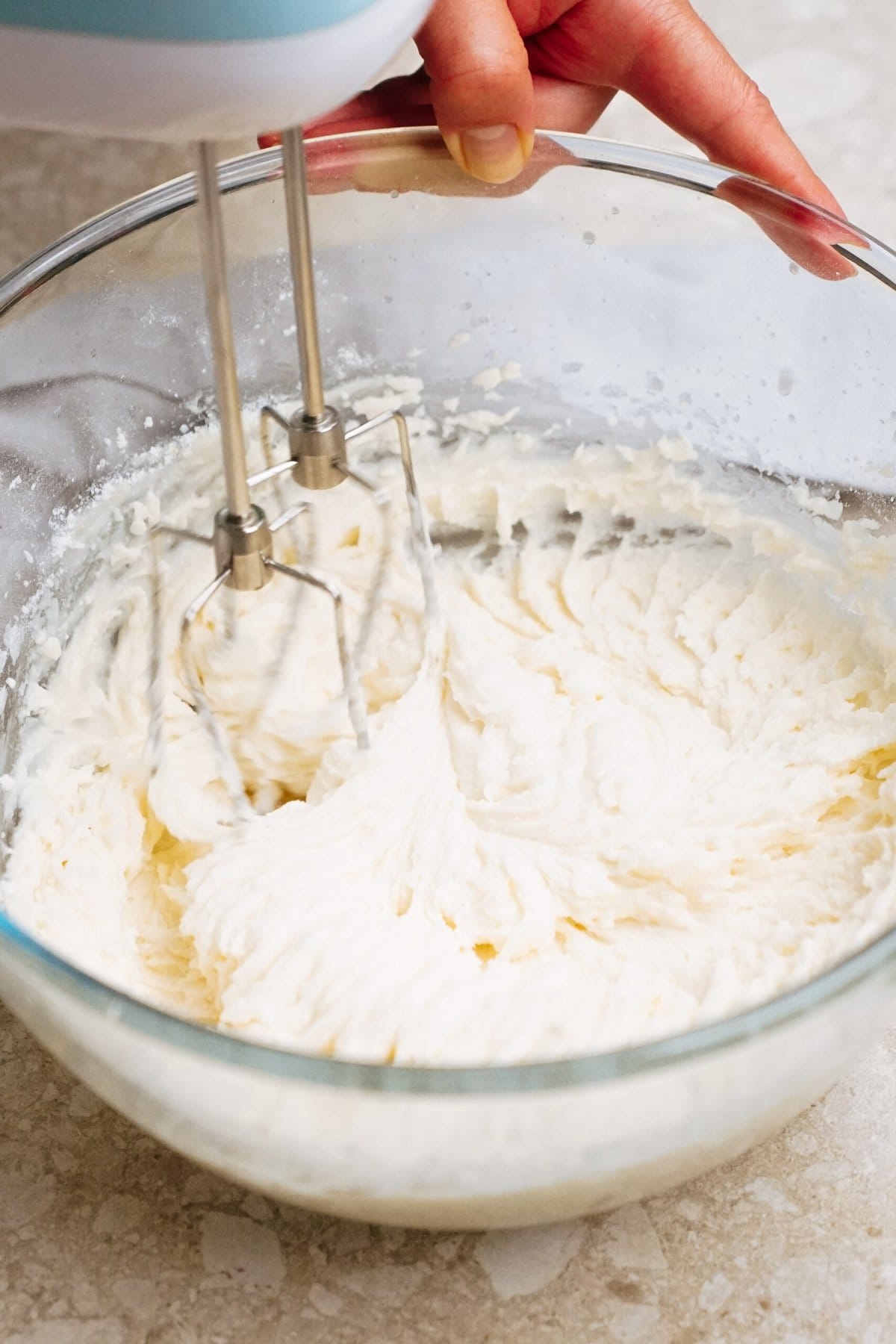 A hand holds a mixer blending a thick, creamy substance in a clear glass bowl.