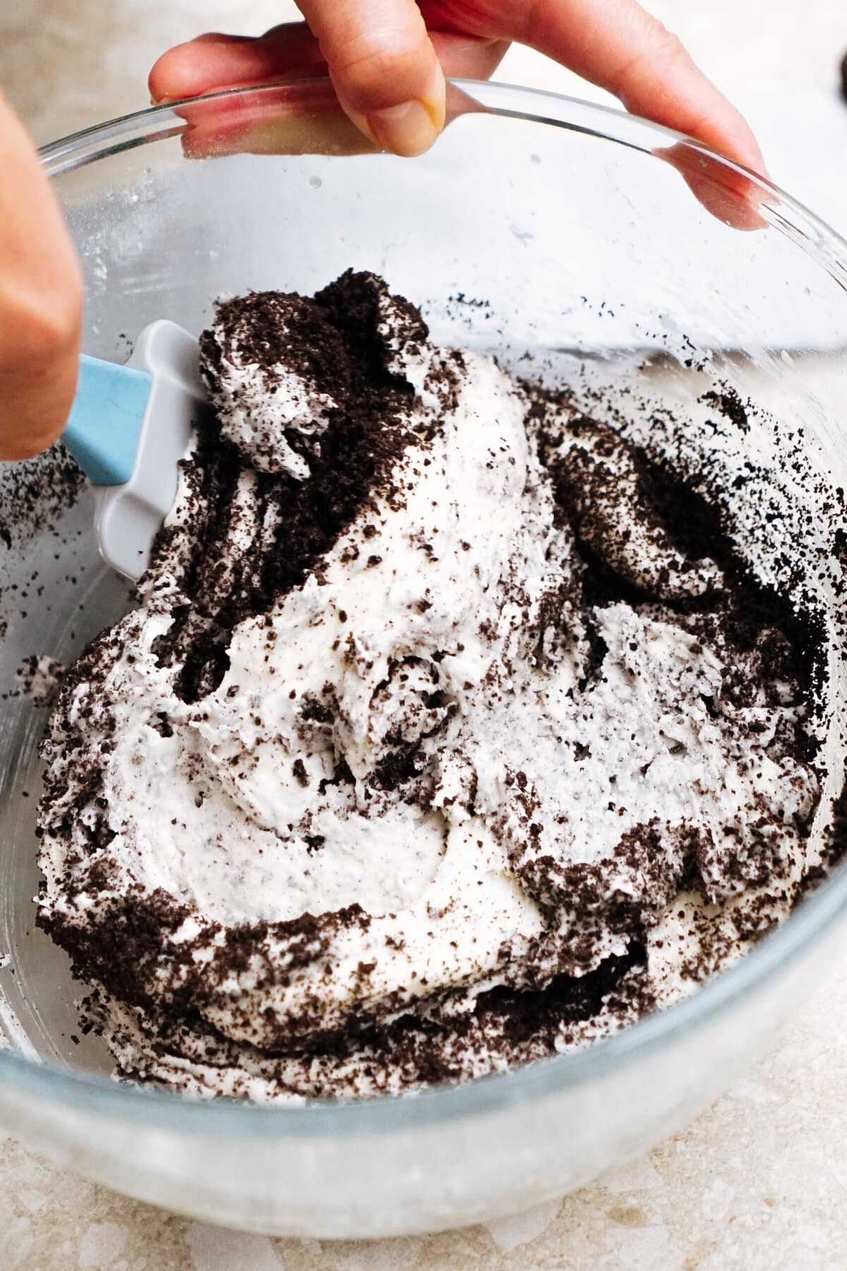 A glass bowl filled with a mixture of crumbled cookies and white creamy substance. A hand is using a spatula to mix the contents.