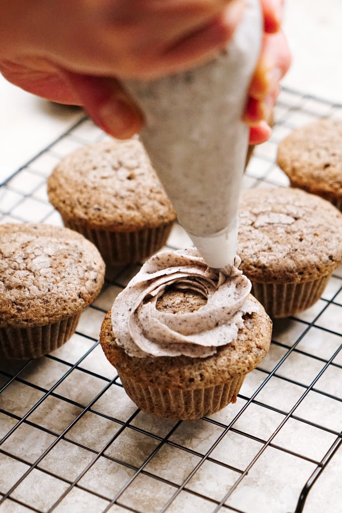 A hand is piping frosting onto a muffin with a piping bag. Several unfrosted muffins are on a cooling rack in the background.
