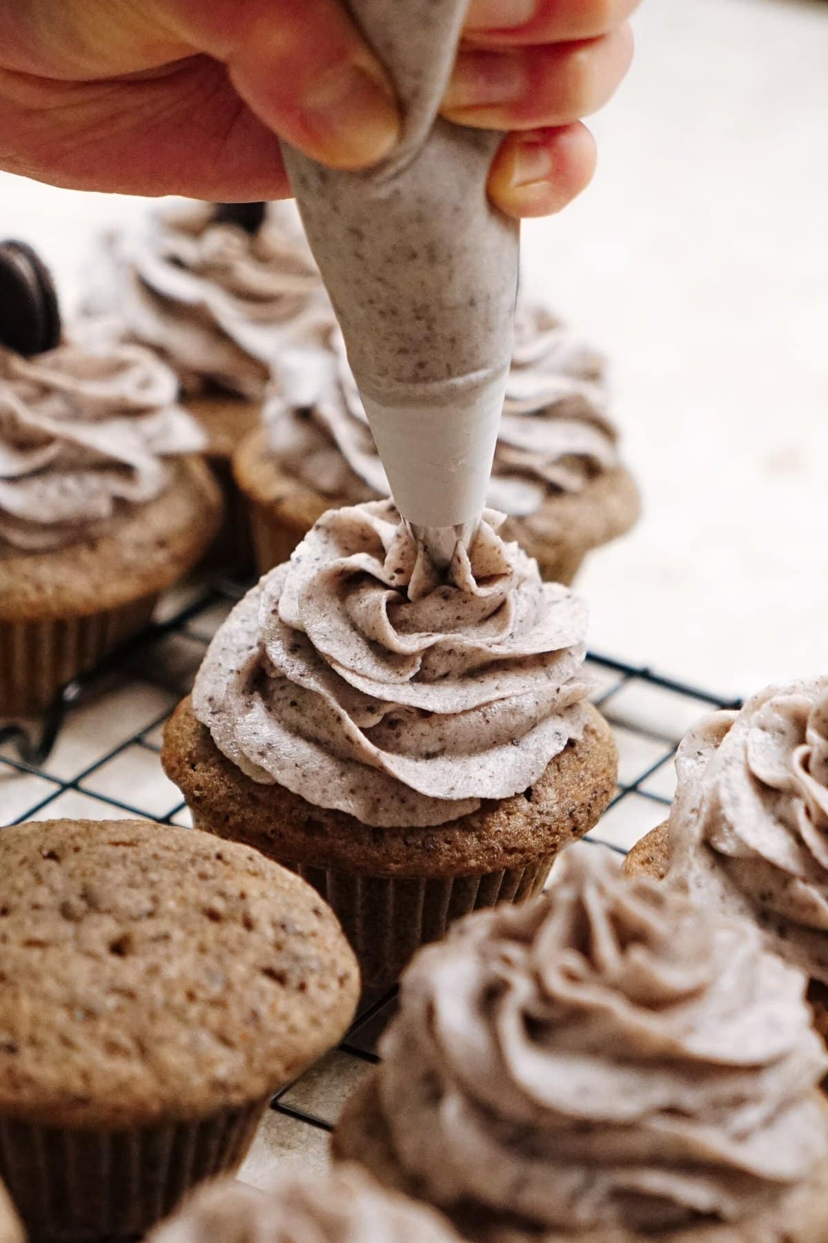 A hand piping chocolate swirled frosting onto cupcakes placed on a cooling rack.