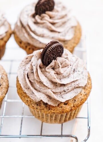 Three cupcakes with swirled cookie and cream frosting, each topped with a mini chocolate sandwich cookie, placed on a cooling rack.