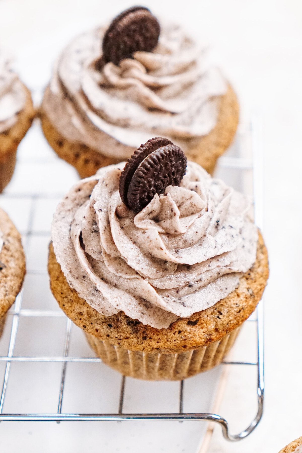 Three cupcakes with swirled cookie and cream frosting, each topped with a mini chocolate sandwich cookie, placed on a cooling rack.