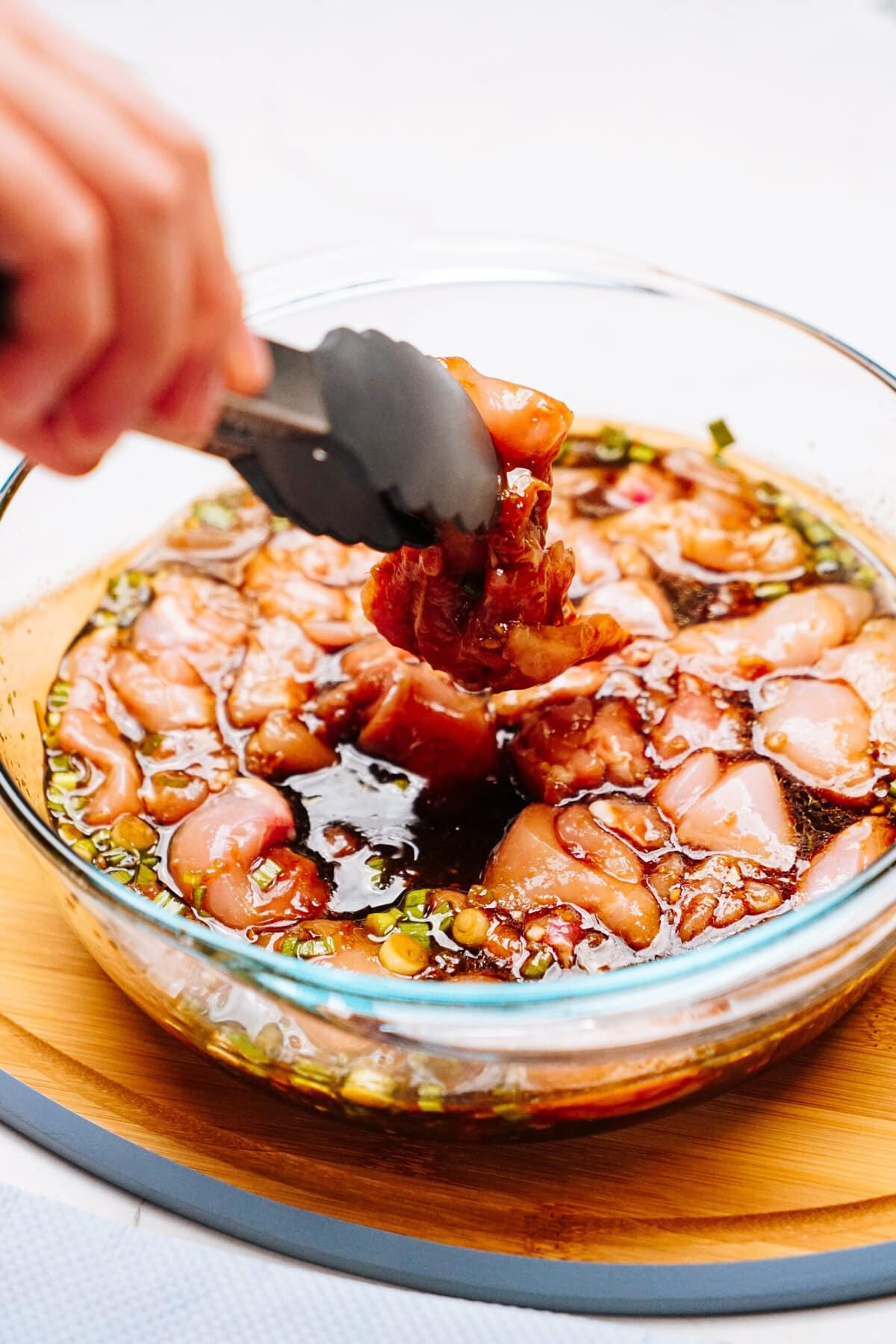 A hand using tongs dips pieces of raw Korean chicken into a marinade in a clear glass bowl.