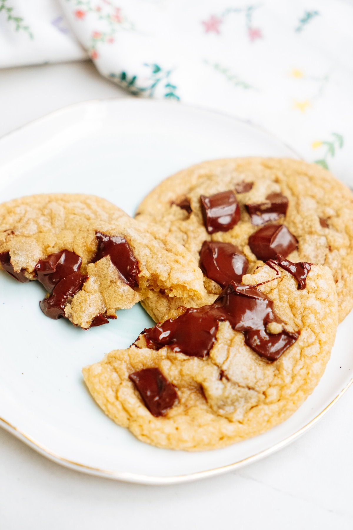 A plate with three chocolate chip cookies, one of which is broken in half, revealing melted chocolate chunks inside. A white cloth with a floral pattern is partially visible in the background.