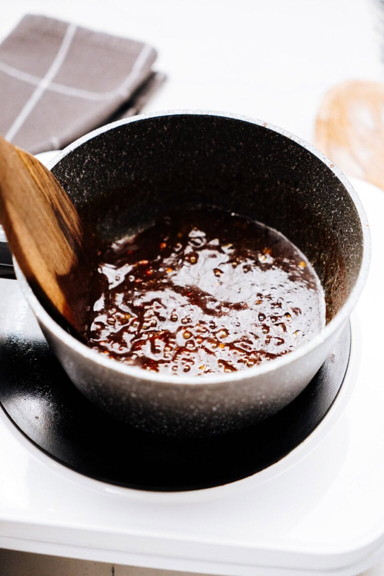 A saucepan containing a thick, simmering brown sauce being stirred with a wooden spoon, reminiscent of the rich flavors found in Korean chicken dishes.