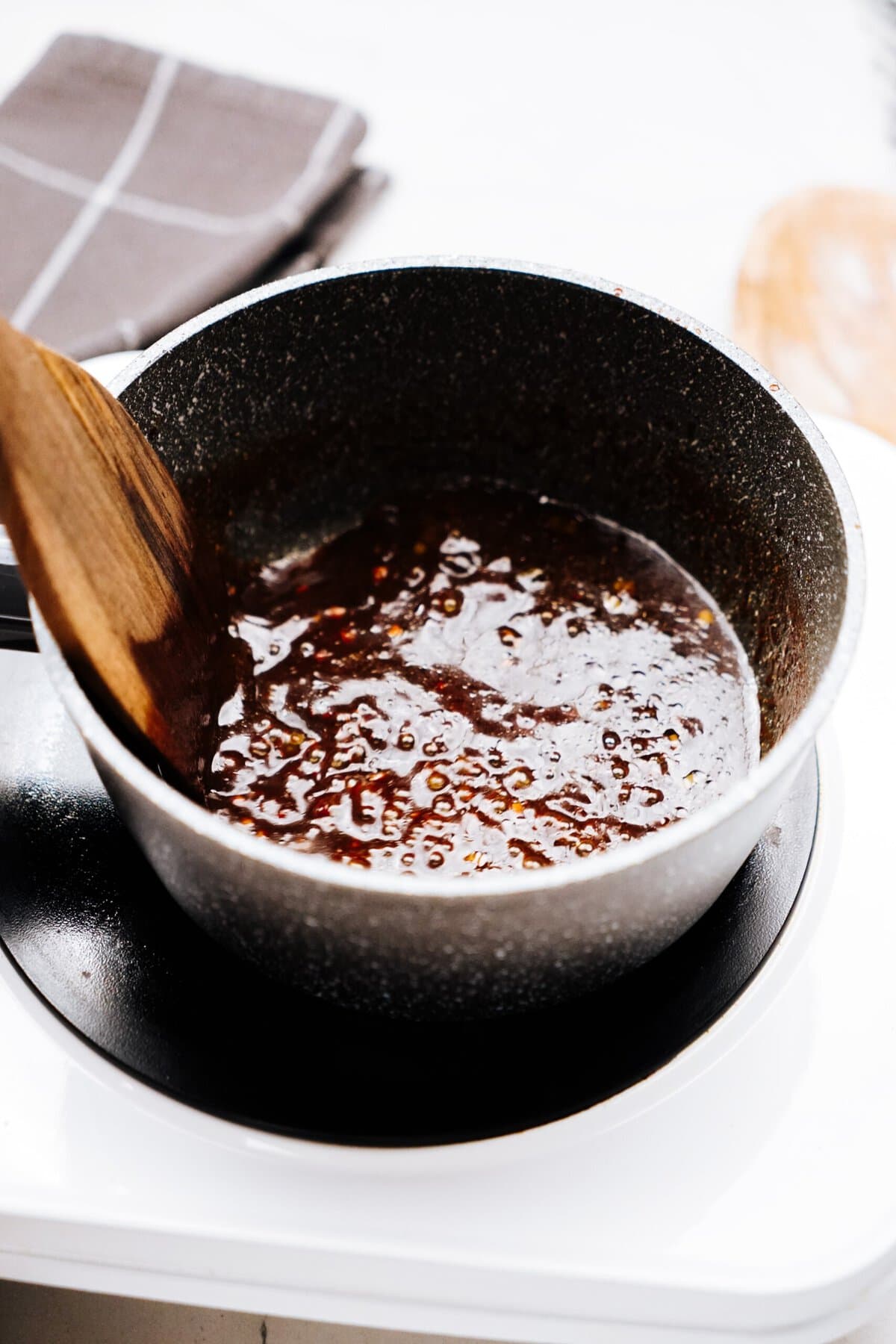 A saucepan containing a thick, simmering brown sauce being stirred with a wooden spoon, reminiscent of the rich flavors found in Korean chicken dishes.