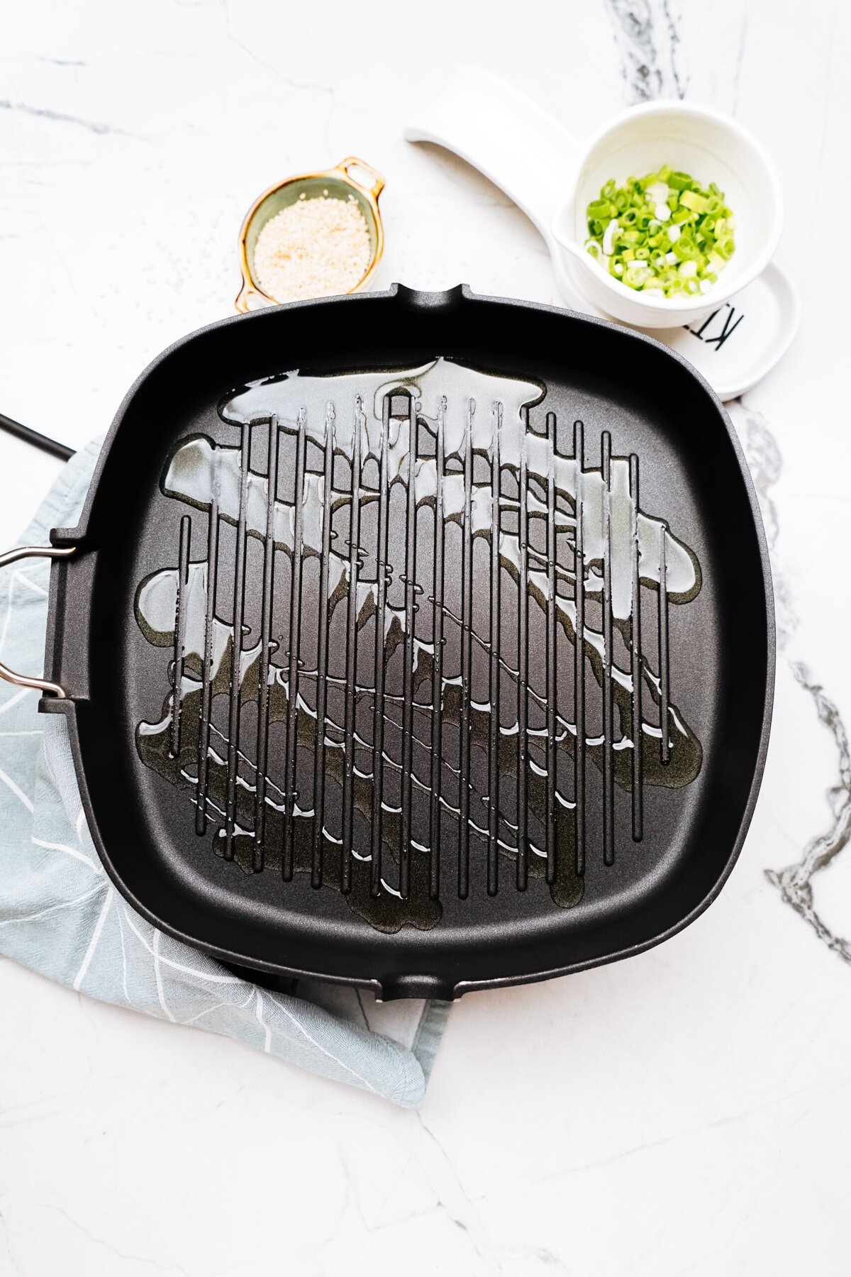 A square grill pan with oil sits on a light marble countertop next to a small bowl of chopped scallions and another small bowl with a yellowish mixture, ready for making Korean chicken. A light blue cloth is underneath the pan's handle.