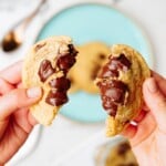 Hands holding a freshly baked chocolate chip cookie split in half, revealing melted chocolate chunks inside, with an aqua plate and another cookie in the background.