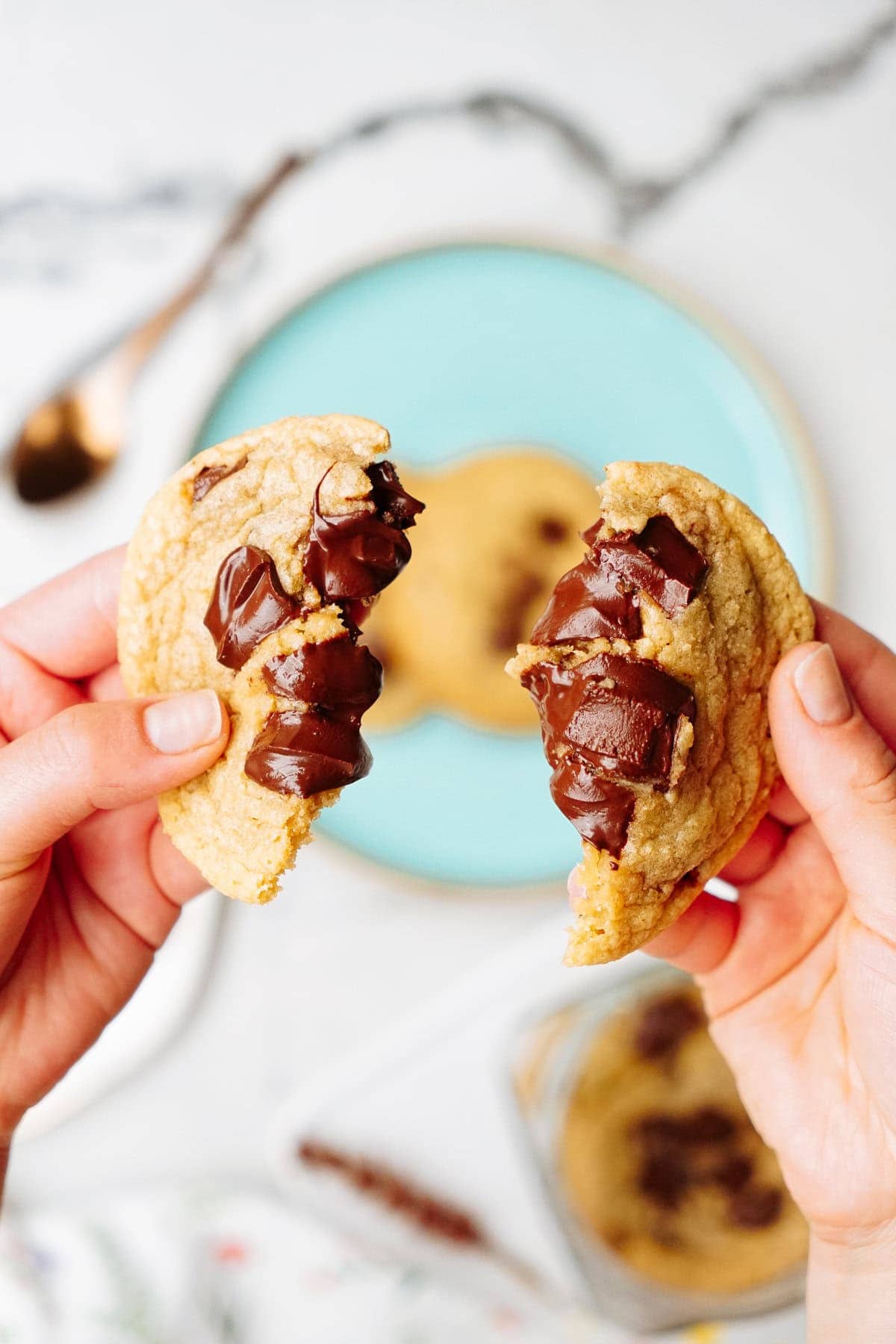 Hands holding a freshly baked chocolate chip cookie split in half, revealing melted chocolate chunks inside, with an aqua plate and another cookie in the background.