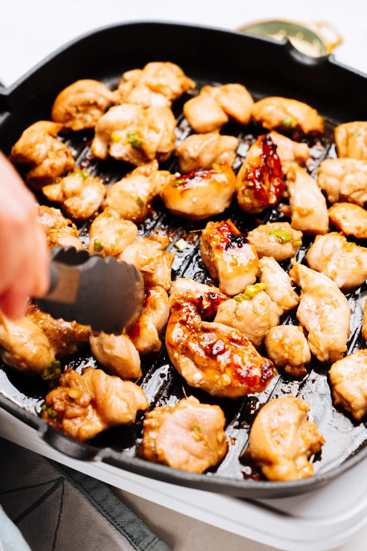 A person uses tongs to cook pieces of marinated Korean chicken on a black grill pan.