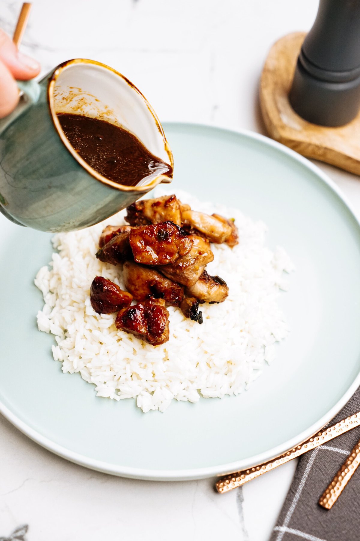 A hand pours sauce from a small jug onto Korean chicken served over white rice on a green plate. Bronze utensils and a gray napkin are placed beside the plate.