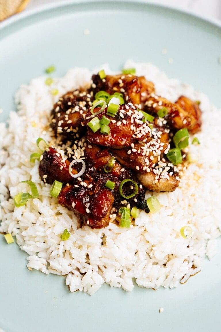 A plate of white rice topped with caramelized Korean chicken pieces, garnished with sliced green onions and sesame seeds.