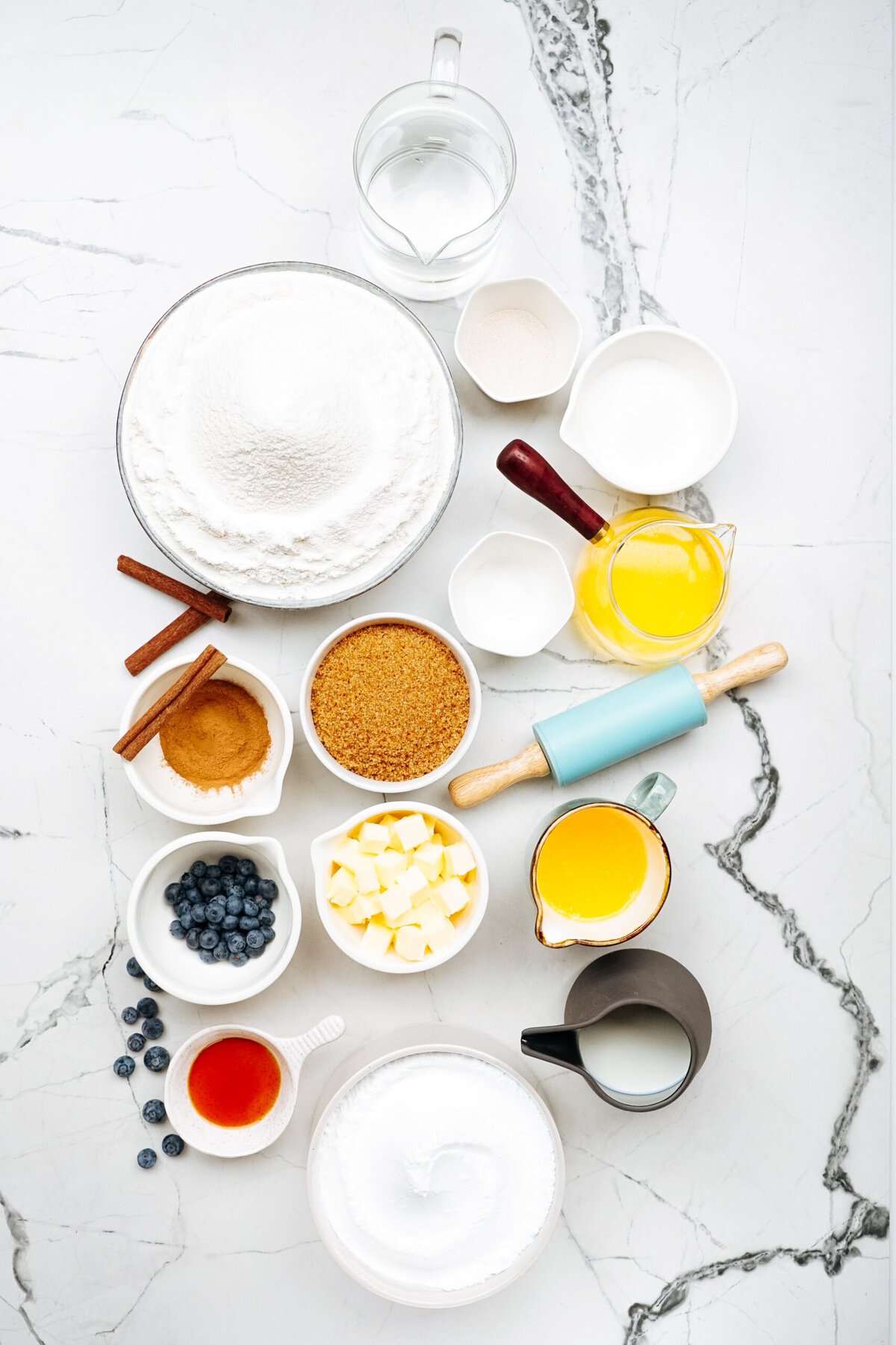 A flat lay of baking ingredients including flour, sugar, butter, eggs, milk, blueberries, cinnamon, brown sugar, and various kitchen tools arranged on a marble surface.