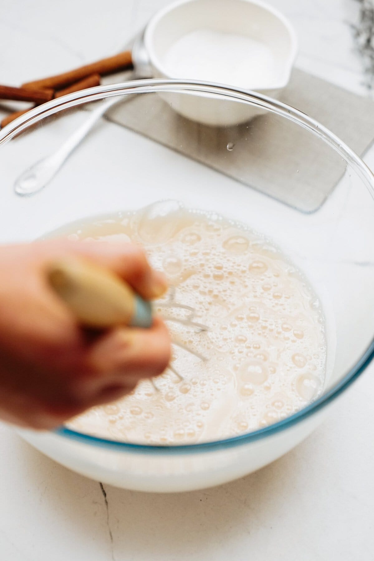 A hand whisking a frothy liquid in a clear glass bowl on a white countertop, with a small white bowl and cinnamon sticks in the background.
