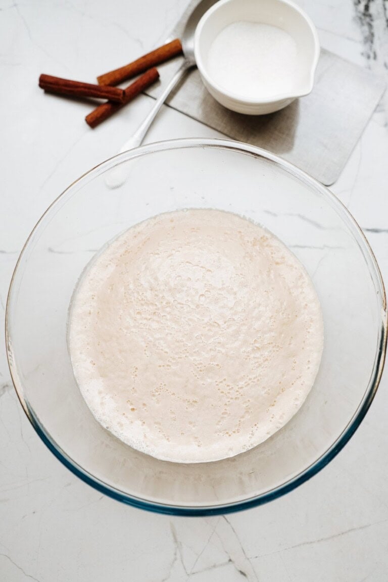 A glass bowl filled with frothy yeast mixture sits on a marble counter, with a small bowl of sugar, a spoon, and cinnamon sticks in the background.