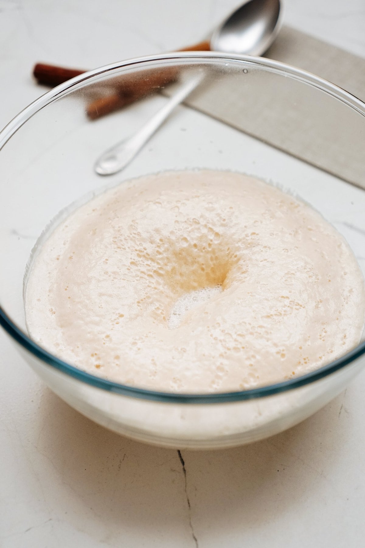 A glass bowl with frothy, activated yeast mixture, with a spoon and kitchen towel in the blurred background on a white surface.