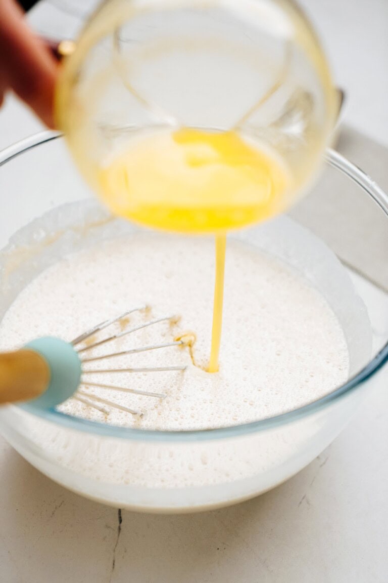 A hand pours a yellow liquid, likely melted butter, into a mixing bowl containing a foamy, white mixture, with a whisk placed inside the bowl.