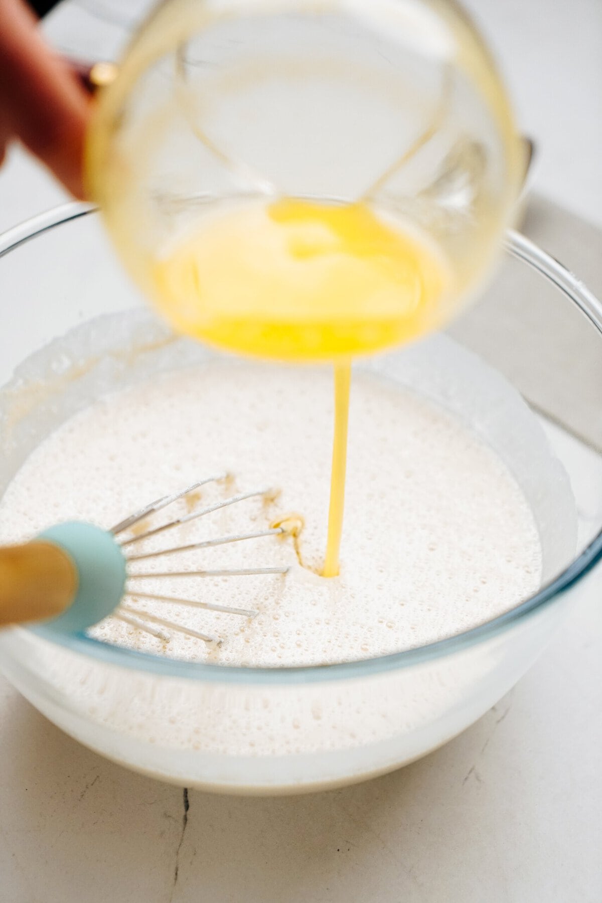 A hand pours a yellow liquid, likely melted butter, into a mixing bowl containing a foamy, white mixture, with a whisk placed inside the bowl.